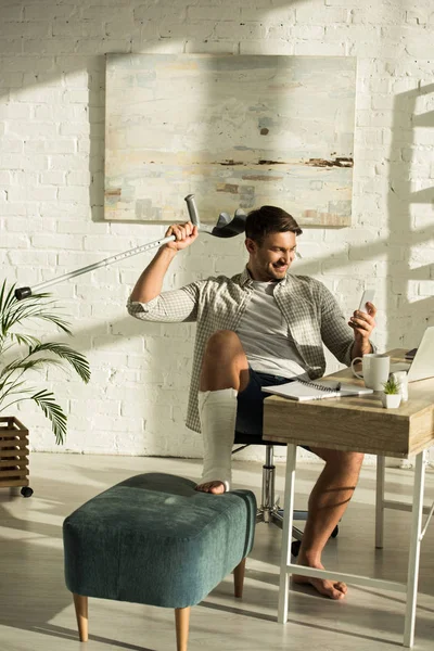 Cheerful freelancer with broken leg holding smartphone and crutch near laptop on table — Stock Photo