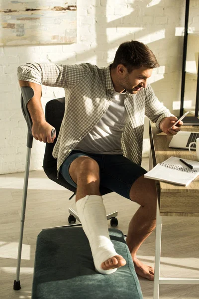 Hombre guapo con pierna en vendaje de yeso usando teléfono inteligente en la mesa con computadora portátil y portátil - foto de stock