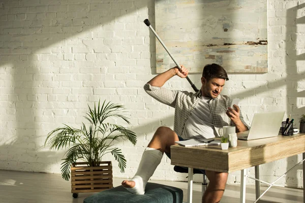 Hombre sonriente con la pierna rota sosteniendo muleta y teléfono inteligente blanco sentado en la mesa con el ordenador portátil - foto de stock