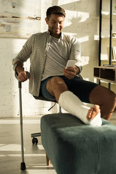 Enfoque selectivo del hombre sonriente con la pierna rota utilizando el teléfono inteligente en la sala de estar — Stock Photo