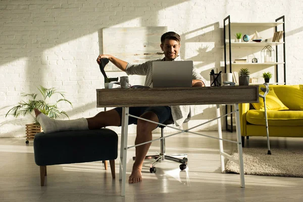 Freelancer sonriente con la pierna rota trabajando en el portátil en la sala de estar - foto de stock