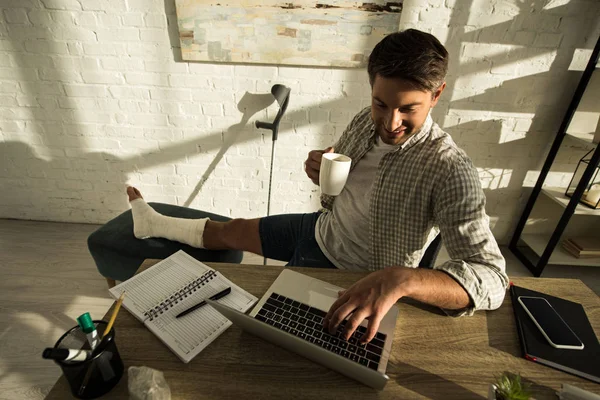 Vista de alto ângulo do freelancer sorridente com perna quebrada segurando copo e trabalhando no laptop na mesa — Fotografia de Stock