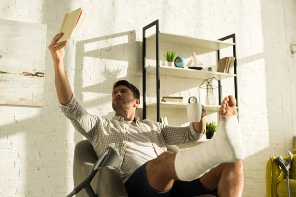 Hombre guapo con la pierna rota levantando libro y sosteniendo la taza mientras está sentado en el sillón en casa - foto de stock