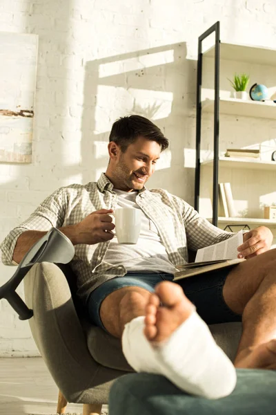 Selektiver Fokus eines lächelnden Mannes mit gebrochenem Bein, der Tasse hält und Buch im heimischen Sessel liest — Stockfoto