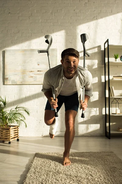 Homme souriant avec une jambe cassée tenant des béquilles à la maison — Photo de stock