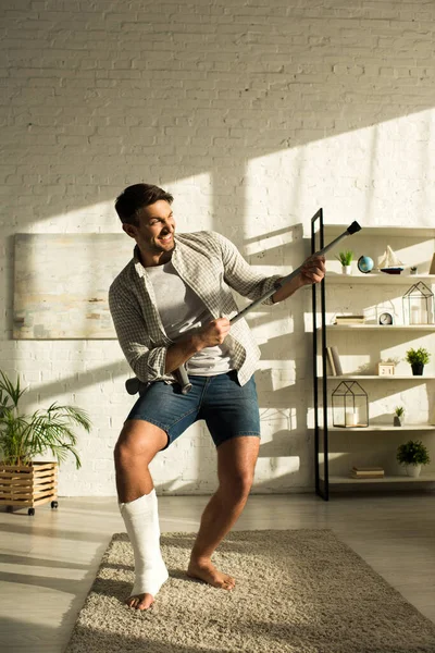 Aufgebrachter Mann mit gebrochenem Bein albert mit Krücke im Wohnzimmer herum — Stockfoto