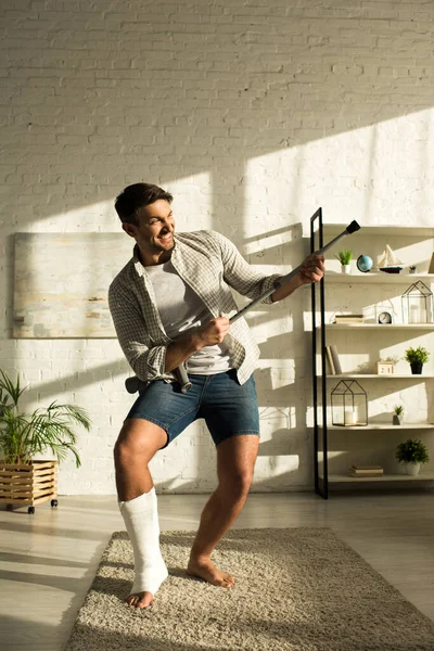 Saído homem com a perna quebrada brincando com muleta na sala de estar — Fotografia de Stock