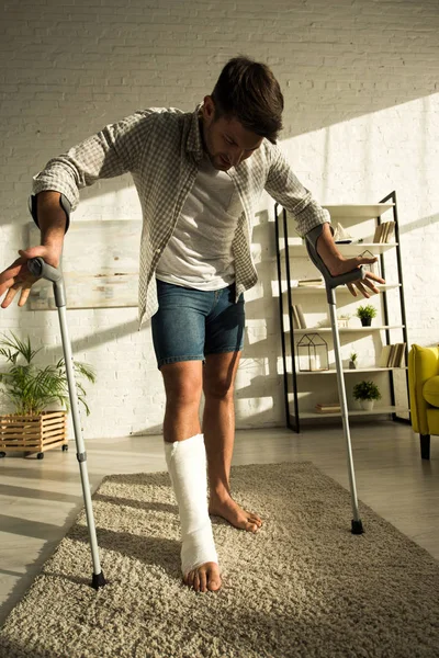 Bonito homem com a perna quebrada segurando muletas enquanto estava na sala de estar — Fotografia de Stock