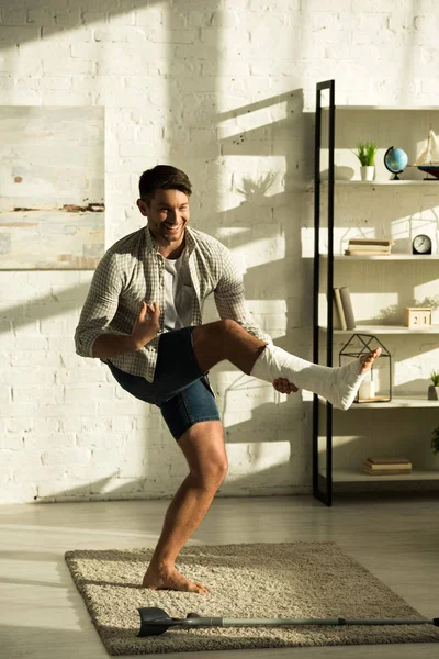 Bello uomo sorridente con la gamba rotta in piedi vicino stampella in soggiorno — Foto stock