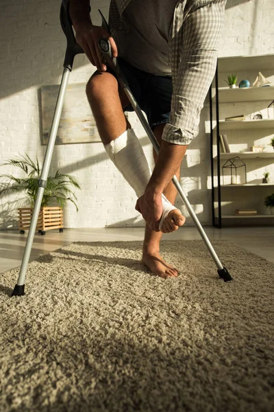 Ausgeschnittene Ansicht von Mann, der gebrochenes Bein berührt und Krücken im Wohnzimmer hält — Stockfoto