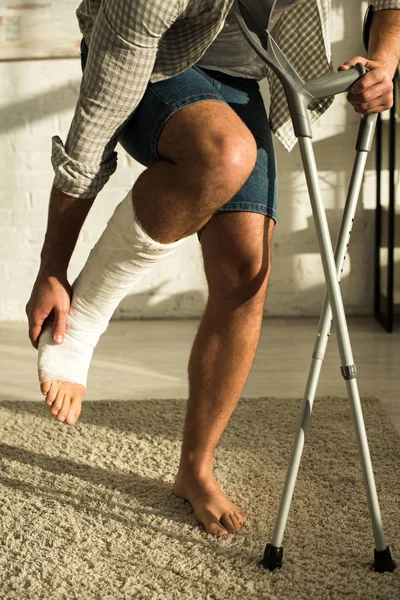 Vista recortada del hombre sosteniendo muletas y tocando la pierna rota en casa - foto de stock