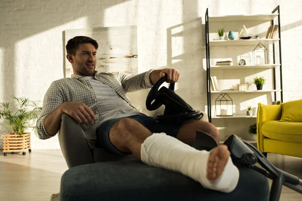 Handsome man with broken leg playing video game with steering wheel in living room — Stock Photo