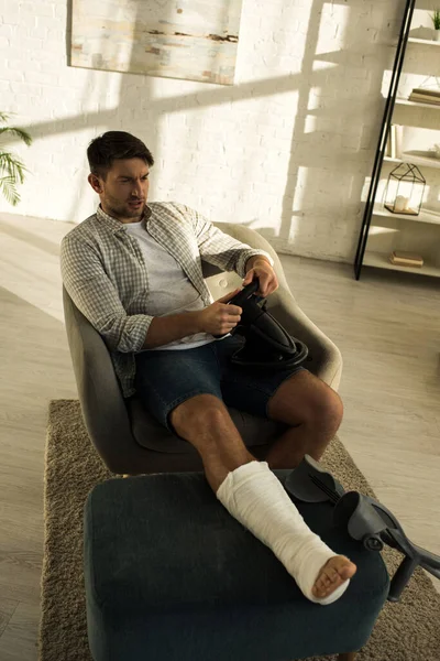 Вид на человека со сломанной ногой под высоким углом, играющего дома в видеоигру с рулевым колесом — стоковое фото