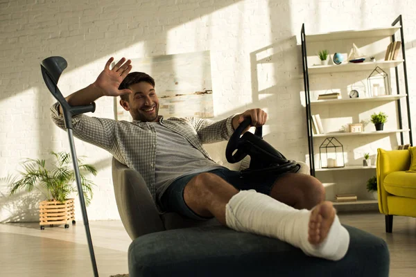 Selective focus of smiling mat with broken leg greeting away while holding playing steering wheel at home — Stock Photo