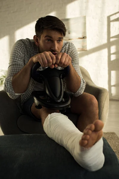 Selective focus of handsome man with broken leg playing video game with steering wheel in armchair — Stock Photo