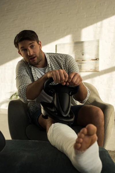 Concentration sélective de l'homme avec la jambe dans le pansement de plâtre tenant le volant de jeu dans le salon — Photo de stock