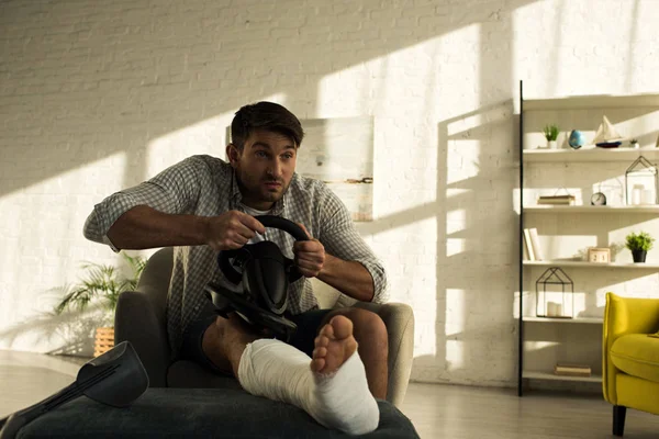 Bell'uomo con la gamba rotta sul pouf che gioca al videogioco con il volante in soggiorno — Foto stock
