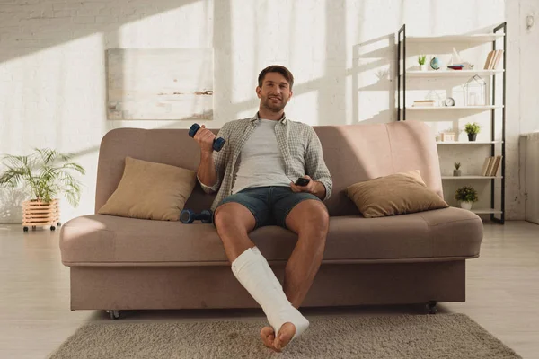 Sorrindo homem com a perna quebrada assistindo tv e segurando halteres no sofá em casa — Fotografia de Stock