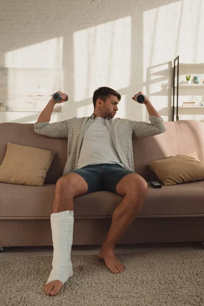 Handsome man with broken leg training with dumbbells on couch in living room — Stock Photo
