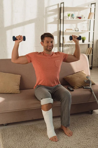 Lächelnder Mann mit gebrochenem Bein trainiert mit Hanteln in der Nähe von Krücken auf der Couch — Stockfoto