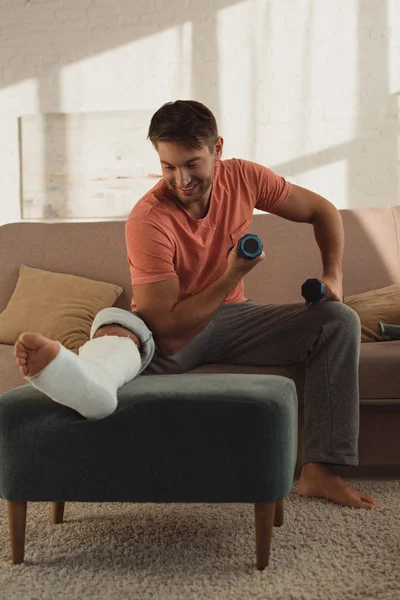 Sorrindo homem com halteres olhando para a perna quebrada no otomano na sala de estar — Fotografia de Stock