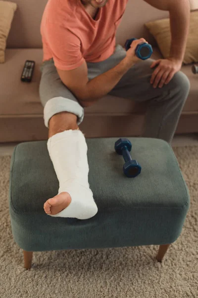 Cropped view of man with broken leg on ottoman working out with dumbbell on couch — Stock Photo