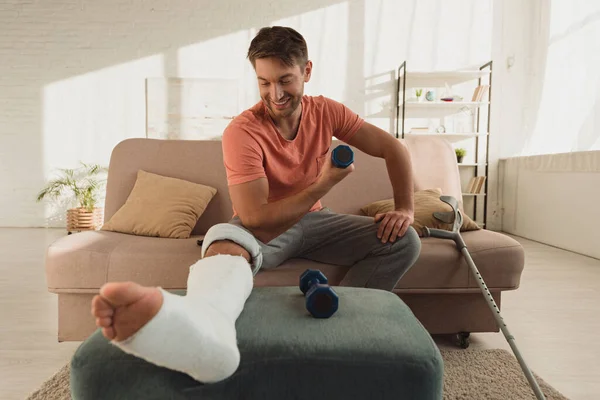 Selective focus of smiling man with broken leg working out with dumbbell on sofa — Stock Photo
