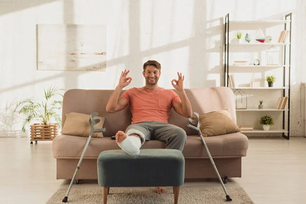 Bello uomo con la gamba rotta sorridente alla fotocamera e mostrando segno ok sul divano — Foto stock