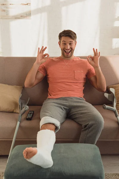 Uscito uomo con gamba rotta sul pouf mostrando segno positivo sul divano a casa — Foto stock