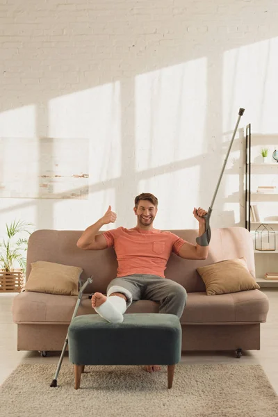 Sorrindo homem com perna quebrada segurando muleta e mostrando o polegar para cima no sofá — Fotografia de Stock