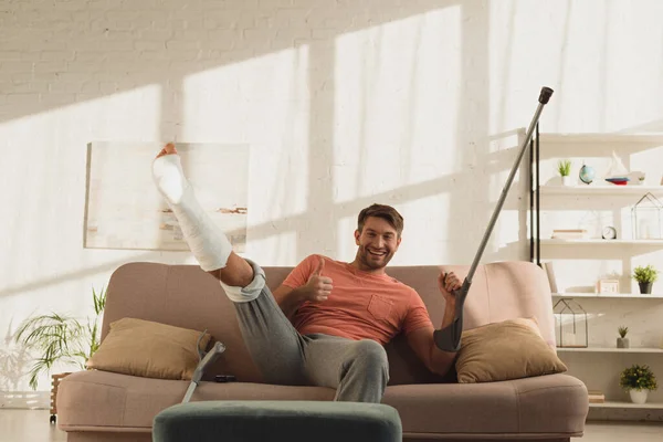 Hombre guapo con la pierna rota levantada y la muleta mostrando el cartel de aprobación en la cámara en el sofá - foto de stock