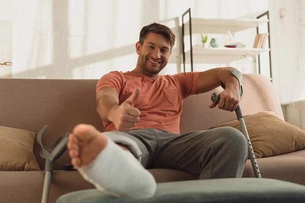 Focus sélectif de l'homme souriant avec jambe cassée montrant le pouce vers le haut symbole sur le canapé à la maison — Photo de stock