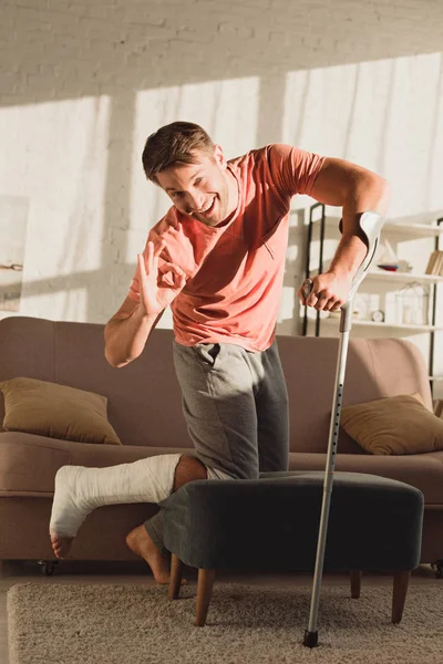 Mann mit gebrochenem Bein lächelt in Kamera und zeigt Okay-Zeichen zu Hause — Stockfoto