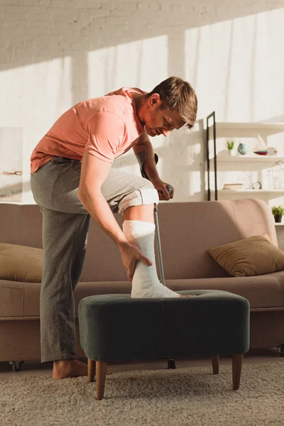 Vue latérale de l'homme touchant jambe cassée sur pouf à la maison — Photo de stock