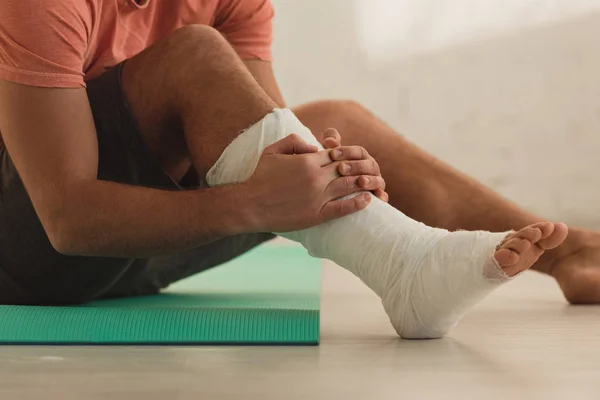 Cropped view of man holding broken leg and sitting on fitness mat on floor — Stock Photo