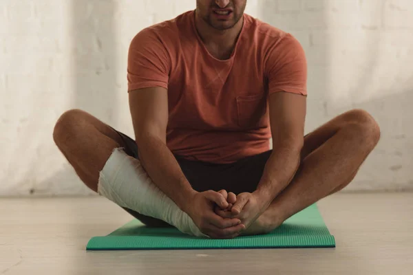 Cropped view of man with broken leg stretching on fitness mat on floor — Stock Photo