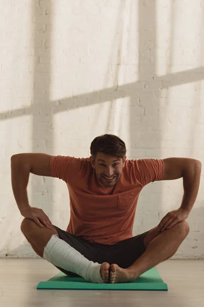 Uomo sorridente con gamba in benda di gesso che guarda la fotocamera e si estende sul tappeto fitness sul pavimento — Foto stock