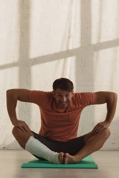 Smiling man with leg in plaster bandage looking at camera and stretching on fitness mat on floor — Stock Photo