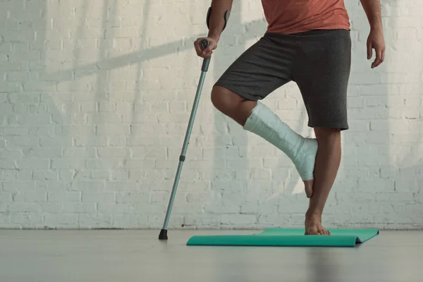 Vue recadrée de l'homme avec jambe cassée debout sur l'homme de remise en forme et tenant la béquille — Photo de stock