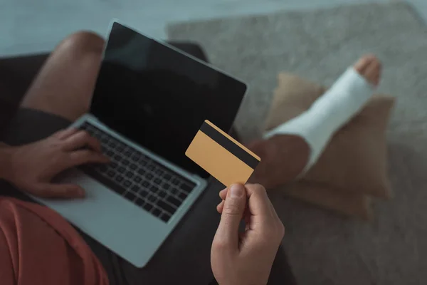 Вид под высоким углом на человека со сломанной ногой, держащего кредитную карту и использующего ноутбук — стоковое фото