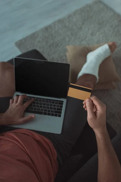 Visão de alto ângulo do homem com a perna em bandagem de gesso usando laptop e segurando cartão de crédito — Fotografia de Stock