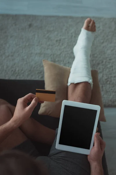 Hochwinkelaufnahme eines Mannes mit Gips am Bein mittels digitalem Tablet und Kreditkarte auf der Couch — Stockfoto