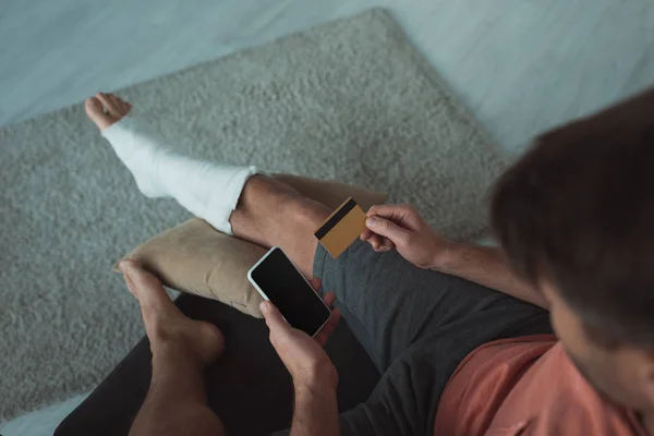 Vue aérienne de l'homme avec jambe cassée en utilisant un smartphone et une carte de crédit sur le canapé — Photo de stock