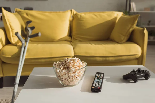 KYIV, UCRAINA - 21 GENNAIO 2020: Focus selettivo di popcorn, telecomando e joystick con stampelle vicino al divano in soggiorno — Foto stock