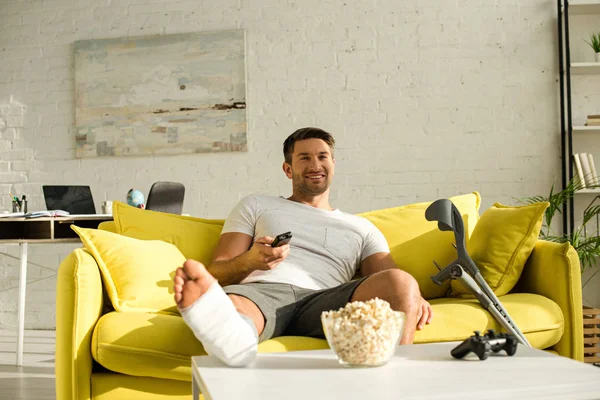 KYIV, UKRAINE - JANUARY 21, 2020: Selective focus of smiling man with broken leg watching tv near popcorn and joystick on coffee table — Stock Photo