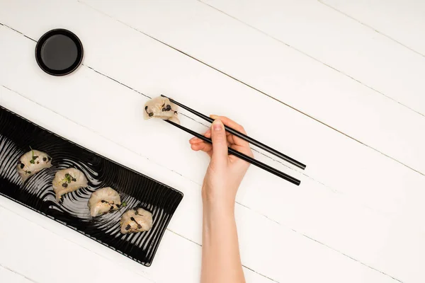 Cropped view of woman eating delicious Chinese boiled dumplings with chopsticks and soy sauce at white wooden table — Stock Photo