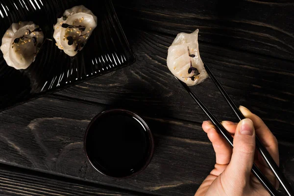 Vue partielle de femme mangeant délicieuse boulette bouillie chinoise avec baguettes et sauce soja à table en bois noir — Photo de stock