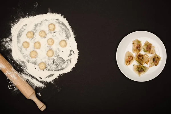 Draufsicht auf köstliche varenyky mit Knistern auf Teller in der Nähe von rohen Teigbällchen, Mehl und Nudelholz auf schwarzem Hintergrund — Stockfoto