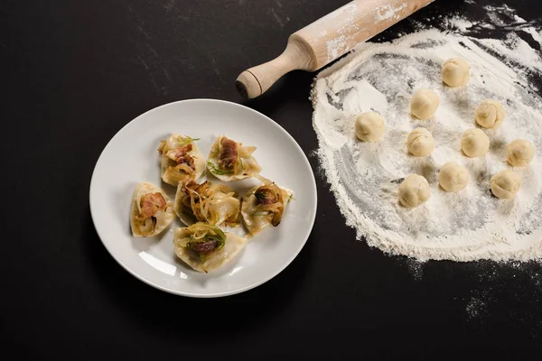 Delicious varenyky with cracklings on plate near raw dough balls, flour and rolling pin on black background — Stock Photo