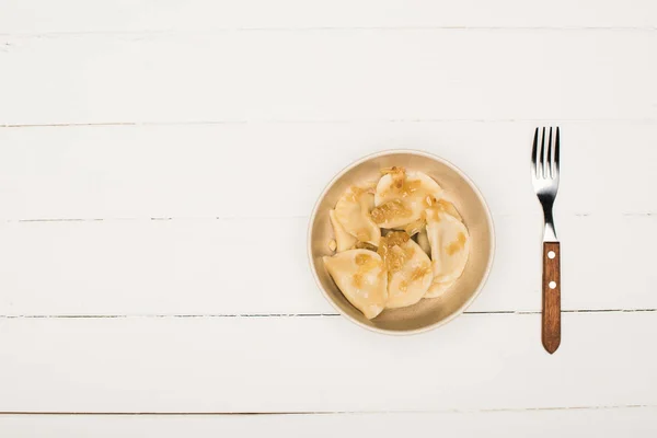 Vue du dessus de délicieux varenyky avec oignon frit dans un bol près de la fourchette sur une table en bois blanc — Photo de stock